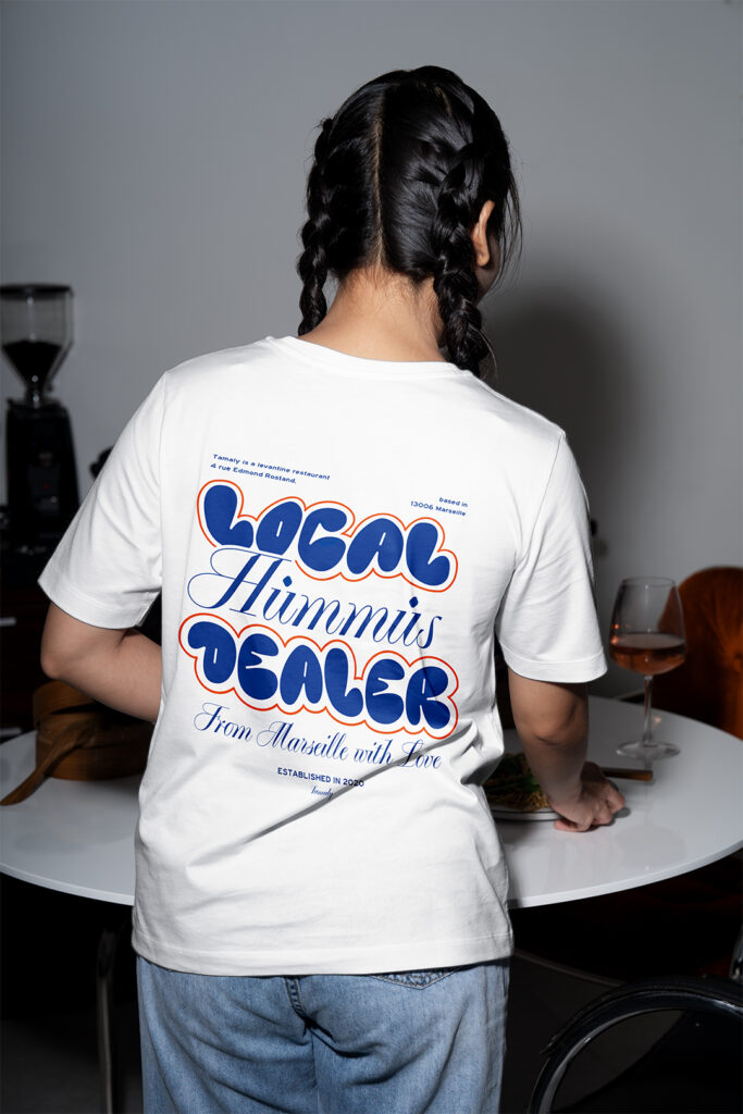 Photographie d'une femme de dos en plein service dans un restaurant qui porte le nouveaux tshirt Tamaly 2025 par John Bertolini