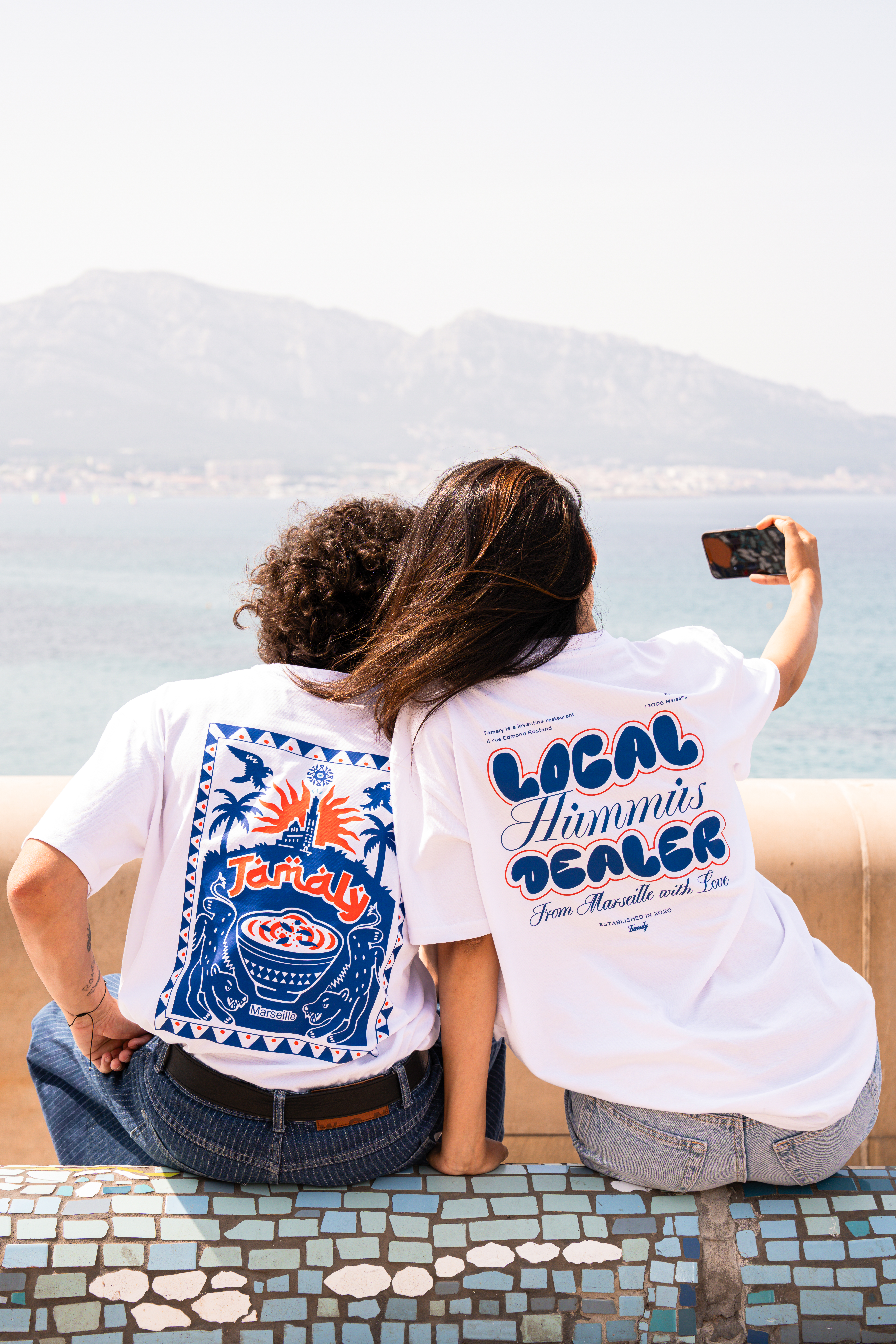 Photographie de deux modèles assis sur un banc face à la mer qui portent les nouveaux tshirt Tamaly 2025 par John Bertolini